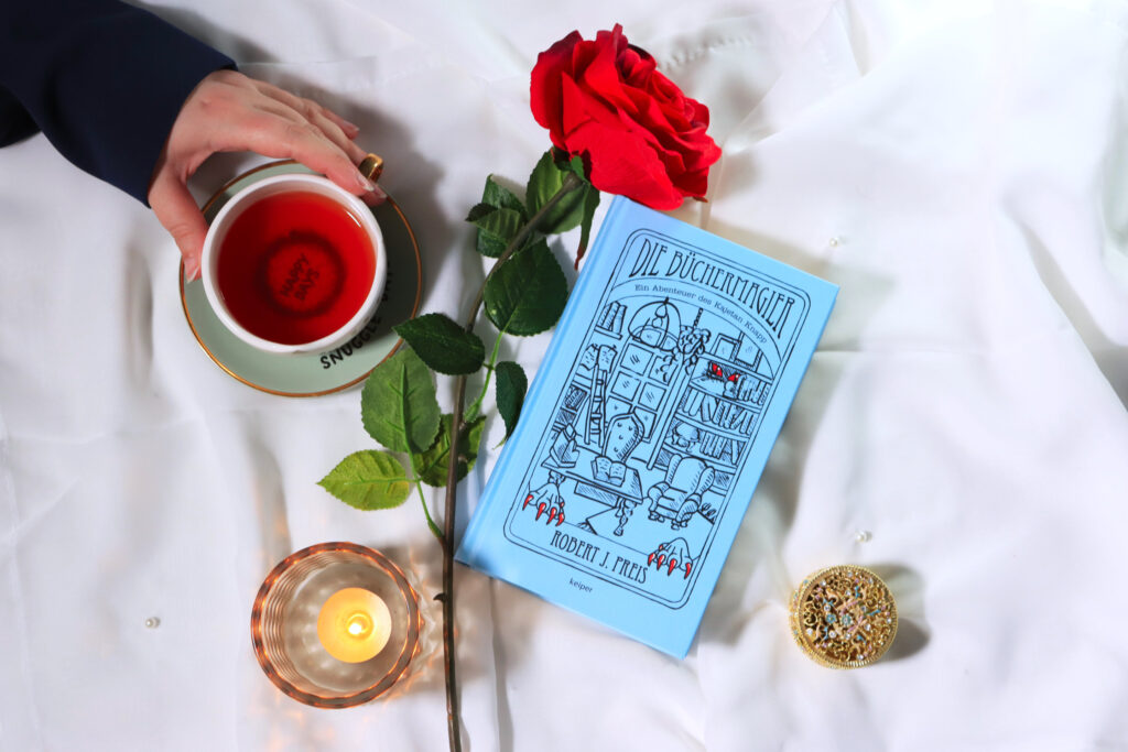 Stillleben mit dem Buch „Die Büchermagier“, roter Rose, Kerze, Schmuckdose und einer Tasse Tee.