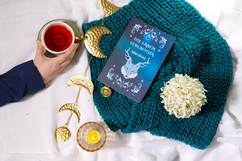 Stillleben mit dem Buch „Die Erben von Botha“, Tasse Tee, goldener Mondgirlande, Blütenkugel, Kerze und gestrickter Decke.