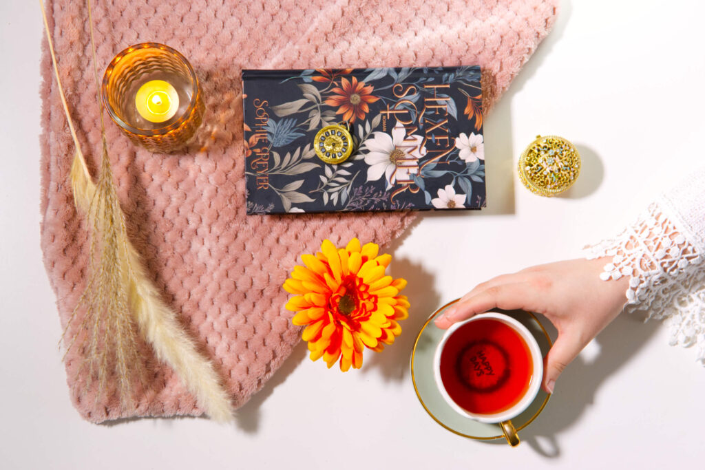 Stillleben mit dem Buch „Hexensommer“, Teetasse, Blume, Kerze, Schmuckdose und rosafarbener Decke.