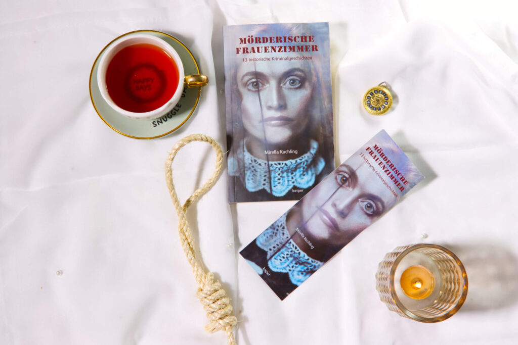 Stillleben mit dem Buch „Mörderische Frauenzimmer“, passendem Lesezeichen, Teetasse, Strickschlinge, Kerze und Schmuckdose auf weißem Stoff.