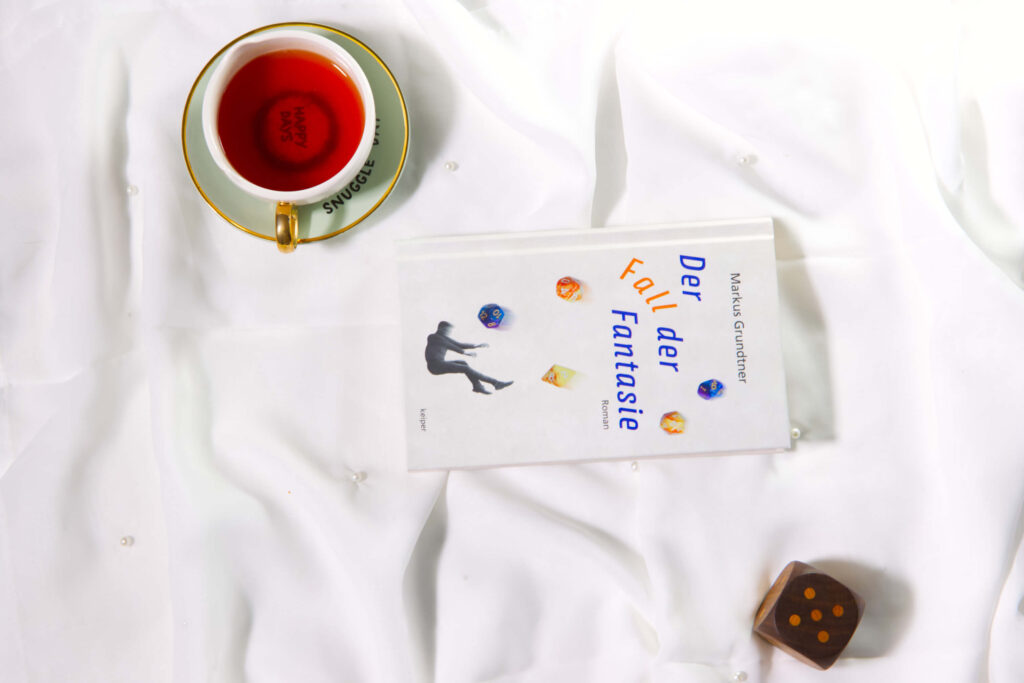 Stillleben mit dem Buch „Der Fall der Fantasie“, Teetasse, bunten Würfeln und Holzwürfel auf weißem Stoff.