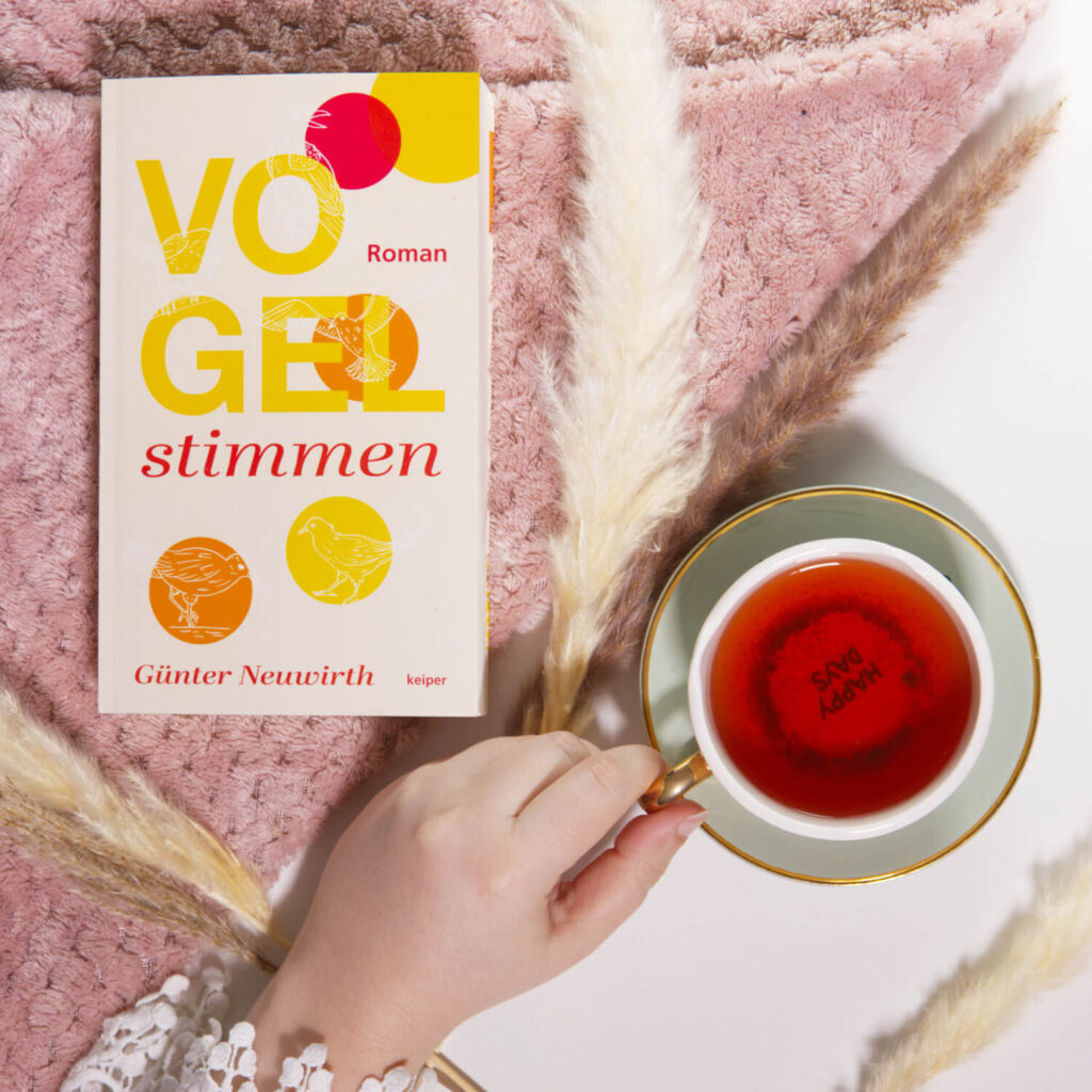 Auf dem mit Pampasgras, einem rosa Tischtuch und einer Teetasse dekorierten Foto wird das Buchcover von "Vogelstimmen" in Szene gesetzt. Das Cover besteht aus verschiedenen Kreisen in Rot, Gelb- und Orange-Tönen. Die Schrift ist prägnant im Cover platziert. Die zarten, weißen Illustrationen verschiedener Vögel runden das Design ab.